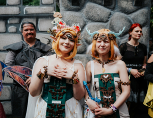 Zwei cosplaying Frauen in fantasievollen Outfits stehen vor einer steinernen Kulisse auf dem Elbenwald Festival. Die Charaktere sind inspiriert von einer fantastischen Welt, umgeben von weiteren Festivalbesuchern in kreativen Kostümen.