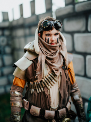 Ein junger Mann in einem detailreichen Steampunk-Kostüm mit Schutzbrille und Kriegsbemalung steht vor einer steinernen Mauer. Sein Outfit ist mit verschiedenen Accessoires und Rüstungsstücken dekoriert, ideal für das Elbenwald Festival.