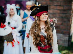 Eine kostümierte Teilnehmerin mit Piratenoutfit und einem Stofftier auf der Schulter, umgeben von anderen Festivalbesuchern in fantasievollen Kostümen. Der Hintergrund zeigt die lebendige Atmosphäre des Elbenwald Festivals.