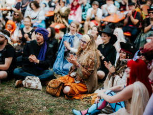 Menschen in fantasievollen Kostümen sitzen auf dem Rasen und applaudieren während einer Veranstaltung im Rahmen des Elbenwald Festivals. Die fröhliche Atmosphäre und die bunte Vielfalt der Besucher spiegeln die kreative Gemeinschaft wider.