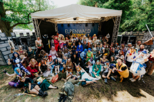 Cosplay Catwalk Gruppenselfie von Festivalbesuchern in aufwendigen Kostümen beim Elbenwald Festival, das Musik und Nerdkultur vereint. Die Teilnehmer posieren fröhlich vor der Bühne, umgeben von thematischen Kulissen und einer lebhaften Atmosphäre.