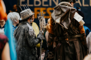 Ein kostümierter Besucher mit einem Skelett-Handschuh und einem auffälligen Hut steht inmitten einer bunten Menschenmenge beim Elbenwald Festival, das Musik und Nerd-Kultur vereint. Im Hintergrund ist das Festival-Logo sichtbar.