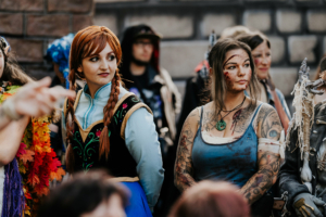 Zwei Frauen in Kostümen auf dem Elbenwald Festival: Eine trägt ein Anna-Kostüm aus "Die Eiskönigin", während die andere mit Tattoos und blutigen Effekten als Charakter erscheint. Im Hintergrund sind weitere Festivalbesucher zu sehen.