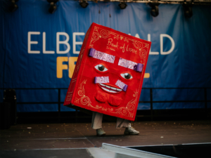 Cosplay Catwalk mit SaKiDe Ein Teilnehmer in einem auffälligen Kostüm als „Buch der Liebe“ auf der Bühne des Elbenwald Festivals, umgeben von einer bunten Kulisse. Die Veranstaltung kombiniert Musik und Nerdkultur und zieht zahlreiche Fans an.