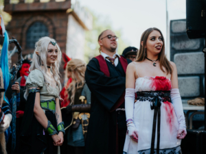 Cosplayer auf dem Elbenwald Festival, gekleidet in fantasievolle Kostüme, darunter eine Elfe mit langen Haaren und eine Figur in einem blutigen Kleid. Im Hintergrund sind weitere Teilnehmer und eine Festivalkulisse zu sehen, die die kreative Atmosphäre unterstreicht.