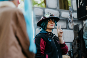 Eine junge Person mit blauen Haaren und einem auffälligen Hut zeigt ein Peace-Zeichen. Sie trägt ein schwarzes Oberteil mit roten Akzenten und posiert vor einer fantasievollen Kulisse. Das Bild fängt die lebendige Atmosphäre des Elbenwald Festivals ein.