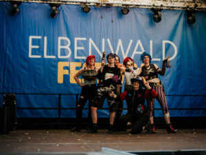 Cosplay Catwalk mit SaKiDe Sechs Cosplayer auf der Bühne des Elbenwald Festivals, gekleidet in fantasievolle Kostüme, posieren fröhlich vor einem großen Bühnenbanner. Die Stimmung ist lebhaft und verspielt, während sie ihre kreativen Outfits präsentieren.