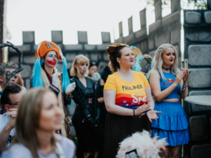 Bunte Festivalbesucher in Kostümen beim Elbenwald Festival, einer Mischung aus Musikfestival und Convention. Die Gruppe besteht aus Menschen mit verschiedenen Outfits, darunter eine clownartige Figur und eine Frau in einem blauen Rock, die applaudiert.