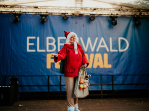 Cosplay Catwalk mit SaKiDe Ein Performer in rotem Mantel und Weihnachtsmütze steht auf der Bühne des Elbenwald Festivals, umgeben von Kuscheltieren. Der Hintergrund zeigt das Festival-Logo, das die kreative und festliche Atmosphäre der Veranstaltung unterstreicht.
