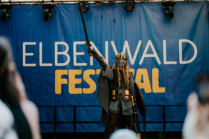 Cosplay Catwalk mit SaKiDe Ein Teilnehmer in beeindruckender Rüstung und mit Schwert auf der Bühne des Elbenwald Festivals, umgeben von einer bunten Menge. Das Festival vereint Musik und Nerd-Kultur und bietet ein einzigartiges Erlebnis für Fans.