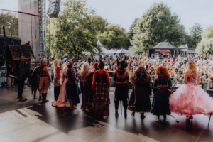 Elbenwald Cosplay Contest Blick auf die Bühne beim Elbenwald Festival mit einer Gruppe von Cosplayern in fantasievollen Kostümen. Im Hintergrund ist eine große Menschenmenge zu sehen, die gespannt auf die Darbietung schaut, umgeben von Bäumen und Festivalzelten.