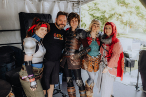 Elbenwald Cosplay Contest Fünf Personen in fantasievollen Kostümen posieren lächelnd hinter der Bühne des Elbenwald Festivals, umgeben von Equipment und einer festlichen Atmosphäre. Die Kostüme zeigen Anklänge an verschiedene Charaktere aus der Popkultur und Fantasy-Welten.