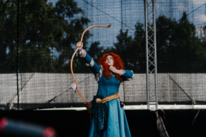 Elbenwald Cosplay Contest Eine cosplayende Person mit roten Locken in einem blauen Kleid zieht einen Bogen auf der Bühne des Elbenwald Festivals, umgeben von einer schwarzen Kulisse. Die Darstellung zeigt die Verbindung von Musik und Fantasiekultur auf dem Event.