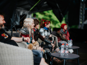 Elbenwald Cosplay Contest Cosplayer auf dem Elbenwald Festival diskutieren auf einer Bühne, umgeben von Wasserflaschen. Die Teilnehmer tragen aufwendige Kostüme, die von verschiedenen Fantasiewelten inspiriert sind. Die Atmosphäre ist lebhaft und kreativ.