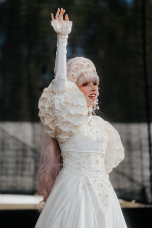 Elbenwald Cosplay Contest Eine Künstlerin in einem eleganten, weißen Kostüm mit Rüschenärmeln und einer verzierten Kopfbedeckung, die während des Elbenwald Festivals auf der Bühne auftritt und das Publikum begeistert.