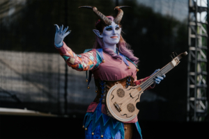 Elbenwald Cosplay Contest Ein fantasievoller Charakter mit blauer Haut und Hörnern steht auf der Bühne des Elbenwald Festivals. Sie trägt ein buntes Kostüm und hält ein einzigartiges Musikinstrument. Die Atmosphäre ist lebhaft und kreativ, perfekt für Fans von Musik und Fantasy.