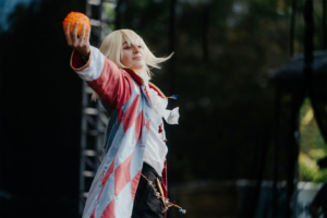 Elbenwald Cosplay Contest Cosplayer auf der Bühne des Elbenwald Festivals, der eine orangefarbene Kugel präsentiert. Im Hintergrund sind Bühnenstrukturen sichtbar, während das Publikum gespannt zuschaut. Die Bildkomposition fängt die lebendige Atmosphäre des Festivals ein.