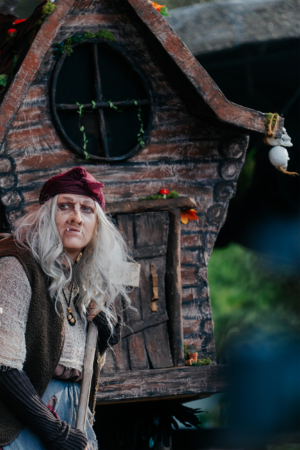 Elbenwald Cosplay Contest Eine Frau in fantasievoller Kostümierung steht vor einem rustikalen, mit Pflanzen dekorierten Holzhaus. Ihre grauen Haare und der Besen verleihen ihr ein mystisches Aussehen, passend zur nerdigen Atmosphäre des Elbenwald Festivals.