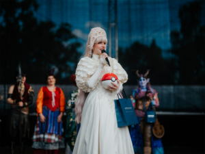 Elbenwald Cosplay Contest Eine Cosplayerin in einem eleganten weißen Kleid hält ein Pokéball-Motiv und spricht ins Mikrofon. Im Hintergrund stehen weitere Teilnehmer in fantasievollen Kostümen. Die Szene zeigt die kreative Atmosphäre des Elbenwald Festivals.