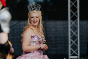 Elbenwald Cosplay Contest Eine fröhliche Teilnehmerin in einem funkelnden, rosa Kostüm mit Krone lächelt auf der Bühne beim Elbenwald Festival, umgeben von anderen Cosplayern. Die Atmosphäre ist lebhaft und kreativ, ideal für Fans von Musik und Popkultur.