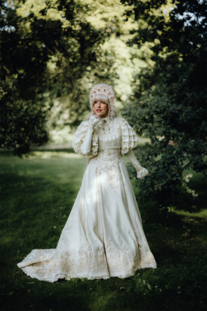 Eine elegant gekleidete Person in einem weißen Kleid mit aufwendigen Verzierungen und einem besonderen Kopfschmuck steht auf einer grünen Wiese. Der Hintergrund zeigt Bäume und eine natürliche Umgebung, die zur Atmosphäre des Elbenwald Festivals passt.