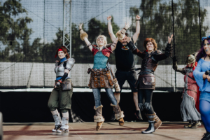 Elbenwald Cosplay Contest Eine Gruppe von Cosplayern in kreativen Kostümen tanzt auf der Bühne beim Elbenwald Festival. Die fröhlichen Teilnehmer zeigen ihre Begeisterung für die Kombination aus Musik und Nerd-Kultur in einer lebhaften Festivalatmosphäre.