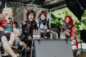 Elbenwald Cosplay Contest Cosplay-Panel auf dem Elbenwald Festival: Vier Teilnehmer in fantasievollen Kostümen sitzen auf der Bühne, während sie sich unterhalten. Im Vordergrund stehen Wasserflaschen, im Hintergrund ist eine grüne Landschaft zu sehen.