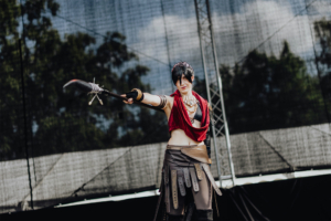 Elbenwald Cosplay Contest Cosplayer auf der Bühne des Elbenwald Festivals, gekleidet in ein auffälliges Kostüm mit rotem Umhang und aufwendigem Zubehör, während er mit einer Waffe posiert. Der Hintergrund zeigt eine Festivalbühne und eine sommerliche Atmosphäre.