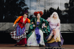 Elbenwald Cosplay Contest Drei Frauen in fantasievollen Kostümen posieren auf der Bühne des Elbenwald Festivals. Die Outfits sind bunt und kreativ, während sie eine lebendige Darbietung zeigen. Im Hintergrund ist eine dunkle Bühnenwand zu sehen, die die Atmosphäre des Festivals unterstreicht.