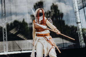 Elbenwald Cosplay Contest Eine cosplayende Person mit roten Haaren, gekleidet in rustikale, fantasievolle Rüstung, steht auf der Bühne des Elbenwald Festivals und hält einen Speer. Im Hintergrund sind Bühnenlichter und eine verschwommene Kulisse zu sehen.
