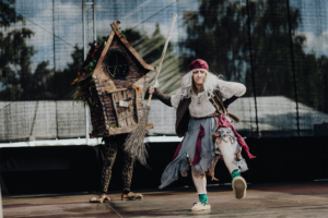 Elbenwald Cosplay Contest Auf der Bühne des Elbenwald Festivals tanzt eine verkleidete Darstellerin in einem zerrissenen Kleid und mit einem Besen, während im Hintergrund eine hölzerne Figur in Form eines Hauses steht. Die lebendige Performance zieht die Aufmerksamkeit der Festivalbesucher auf sich.