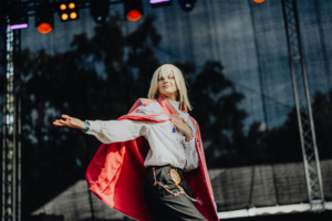 Elbenwald Cosplay Contest Cosplayer in einem auffälligen Kostüm mit rotem Umhang auf der Bühne des Elbenwald Festivals, umgeben von bunten Lichtern. Die Person posiert selbstbewusst und bringt die kreative Atmosphäre des Festivals zum Ausdruck.