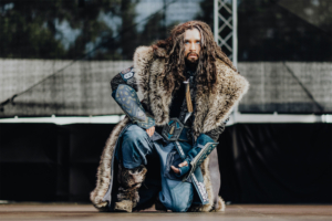 Elbenwald Cosplay Contest Ein kostümierter Teilnehmer des Elbenwald Festivals kniet auf der Bühne, gekleidet in ein aufwändiges Outfit mit pelzbesetztem Umhang und detailreichen Rüstungsteilen. Die Szenerie fängt die kreative Atmosphäre des Festivals ein, das Musik und Nerd-Kultur vereint.