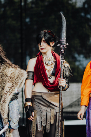 Elbenwald Cosplay Contest Cosplayer auf der Bühne des Elbenwald Festivals, gekleidet in ein detailreiches Kostüm mit rotem Schal und einem Speer. Die Atmosphäre ist lebhaft, umgeben von anderen Teilnehmern in kreativen Outfits, die die Verbindung von Musik und Nerdkultur feiern.