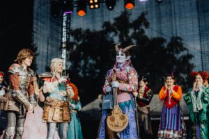 Elbenwald Cosplay Contest Cosplayer auf der Bühne des Elbenwald Festivals, gekleidet in aufwendige Kostüme, während ein Charakter mit einer Laute im Mittelpunkt steht. Im Hintergrund applaudieren andere Teilnehmer, die die kreative Atmosphäre des Festivals unterstreichen.