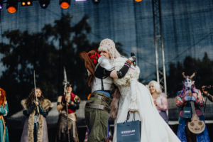 Elbenwald Cosplay Contest Zwei Cosplayerinnen umarmen sich auf der Bühne des Elbenwald Festivals, umgeben von weiteren verkleideten Teilnehmern. Die Atmosphäre ist festlich und voller Begeisterung, während die Zuschauer die kreative Vielfalt der Kostüme genießen.