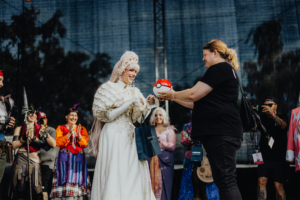 Elbenwald Cosplay Contest Eine Teilnehmerin im aufwendigen Kostüm erhält eine Pokéball-Überraschung auf der Bühne des Elbenwald Festivals, umgeben von jubelnden Fans in fantasievollen Outfits. Die Atmosphäre ist festlich und kreativ, perfekt für Fans von Popkultur und Musik.