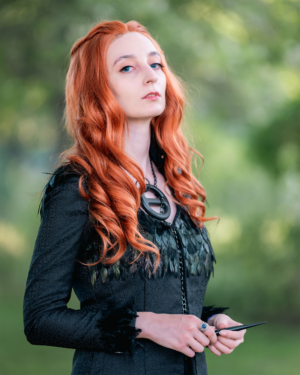 Kostümierte Teilnehmerin mit langen, roten Haaren und schwarzem Outfit mit Federdetails, die beim Elbenwald Festival in der Natur posiert. Sie hält einen Stift in der Hand und strahlt eine mystische Aura aus, umgeben von grüner Kulisse.
