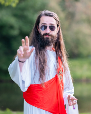 Ein langhaariger Mann in Sonnenbrille und weißem Gewand mit rotem Umhang posiert lässig am Elbenwald Festival, das Musik und Nerdkultur vereint. Seine entspannte Haltung und das freundliche Lächeln spiegeln die kreative Atmosphäre des Festivals wider.