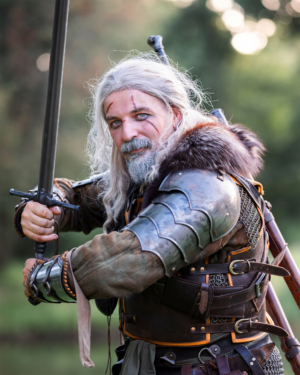 Ein kostümierter Teilnehmer des Elbenwald Festivals, gekleidet als fiktiver Charakter mit langem grauen Haar, Schwert in der Hand und detaillierter Rüstung. Die Szene zeigt eine lebendige Verbindung von Fantasy und Musik in einer grünen Umgebung.