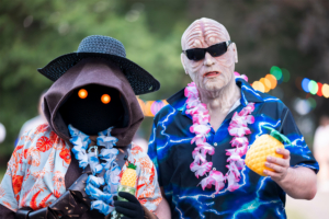 Zwei Festivalbesucher in kreativen Kostümen beim Elbenwald Festival, einer Mischung aus Musikfestival und Convention. Der erste trägt ein tropisches Hemd und Sonnenhut, der zweite hat eine auffällige Maske und Sonnenbrille, beide halten Getränke in Hand.