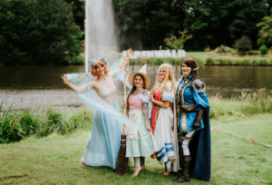 Vier Frauen in fantasievollen Kostümen stehen am Ufer eines Teichs beim Elbenwald Festival. Sie posieren fröhlich vor dem Festival-Schild, umgeben von grüner Natur und einem Wasserfall im Hintergrund. Die Kostüme sind inspiriert von Märchen und Fantasy.
