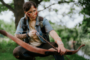 Eine Person mit Tattoos und Verletzungen zielt mit einem Bogen auf dem Elbenwald Festival. Die Szene zeigt eine Mischung aus Cosplay und Abenteuer in einer grünen Umgebung, die die kreative Atmosphäre des Festivals unterstreicht.