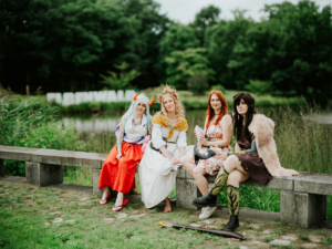 Cosplay-Gruppenfoto beim Elbenwald Festival, vier Frauen in fantasievollen Kostümen sitzen auf einer Steinbank vor einem Gewässer. Hinter ihnen ist das Festival-Logo sichtbar, umgeben von üppiger Natur. Die Atmosphäre strahlt Freude und Kreativität aus.