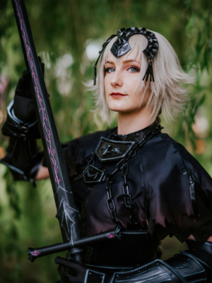 Cosplayer auf dem Elbenwald Festival, gekleidet in ein detailliertes, schwarzes Kostüm mit einer auffälligen Waffe. Der Hintergrund zeigt eine grüne Kulisse, die zur Atmosphäre des Festivals beiträgt. Ideal für Fans von Fantasy und Popkultur.