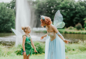 Zwei verkleidete Feen lachen und interagieren am Elbenwald Festival, umgeben von Natur und einem Wasserfall im Hintergrund. Die Szene fängt die magische Atmosphäre des Festivals ein, das Musik und Fantasie vereint.