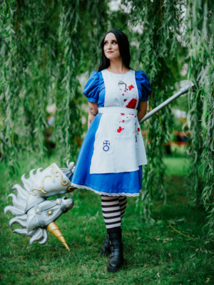 Eine cosplaying Frau in einem blauen Kleid mit blutigen Flecken und gestreiften Strumpfhosen hält einen einzigartigen Waffe in Form eines Einhorns. Der Hintergrund zeigt eine grüne, bewaldete Umgebung, die die Atmosphäre des Elbenwald Festivals unterstreicht.