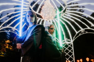 Zwei Cosplayer in beeindruckenden Star-Wars-Kostümen halten leuchtende Lichtschwerter. Die Szenerie ist festlich mit Lichtern dekoriert, während der Elbenwald Festival-Vibe die magische Atmosphäre unterstreicht.
