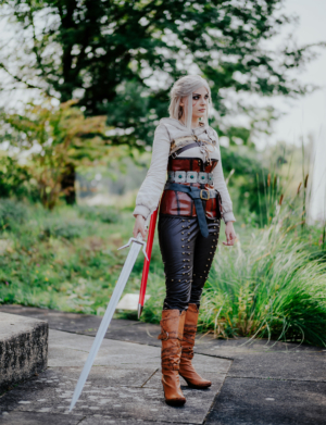 Cosplayer in detailreicher Rüstung mit Schwert auf dem Elbenwald Festival. Die Person steht in einer natürlichen Umgebung, umgeben von Pflanzen, und trägt braune Stiefel sowie eine Kombination aus Leder und Stoff, die an ein Fantasy-Charakterdesign erinnert.