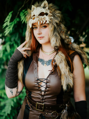 Frau in einem kreativen Kostüm mit Wolfsmütze und Tätowierungen, posiert vor grünem Hintergrund beim Elbenwald Festival. Die Mischung aus Fantasy und Musik spiegelt den einzigartigen Stil und die Atmosphäre des Events wider.